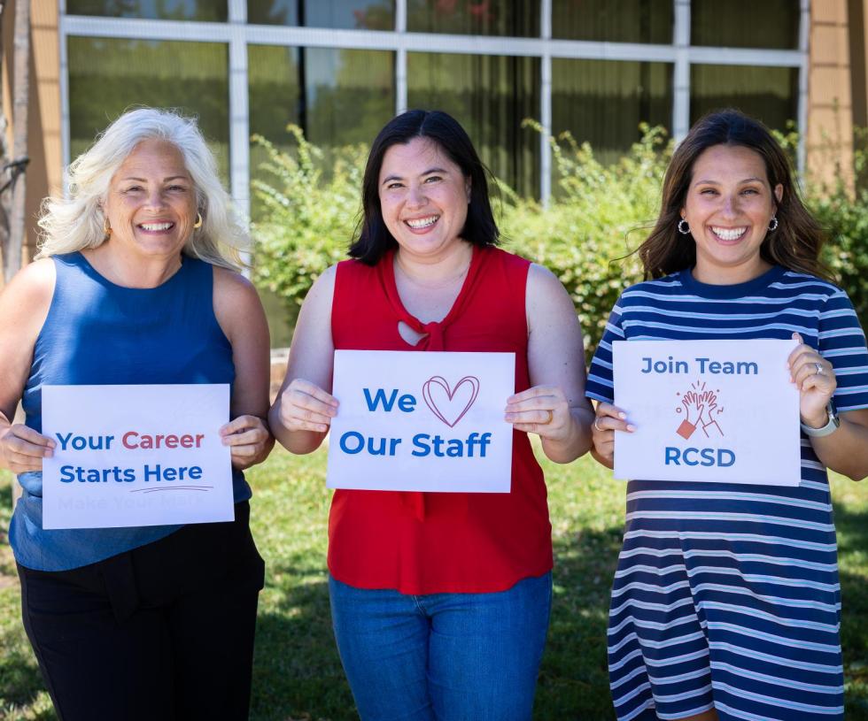 Three employees in front of the District Office, holding signs that say "your career starts here," "We love our staff," and "Join team RCSD"