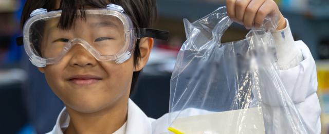 Elementary School student dressed in a lab coat and goggles stands in a library, holding up a plastic bag of school supplies.
